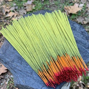 May include: A bundle of incense sticks with green shafts and orange and red tips rests on a dark gray stone. The incense is arranged in a fan shape, with the tips of the sticks fanning out. The background is a natural setting with leaves and grass.
