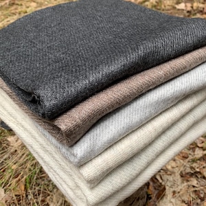 May include: A stack of folded fabric in various neutral colors, including charcoal gray, taupe, and cream. The fabrics have a textured appearance and are arranged on a natural stone surface, surrounded by fallen leaves and foliage.