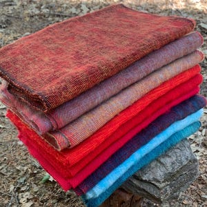 May include: A stack of colorful woven textiles in shades of red, orange, purple, and blue. The textiles are neatly folded and stacked on top of each other, showcasing a variety of textures and hues. The stack is resting on a small wooden stump, with a natural outdoor background.