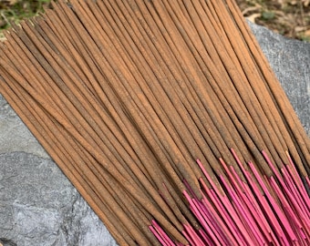 Peut inclure: Un paquet de bâtonnets d'encens bruns avec des pointes roses. Les bâtonnets sont disposés en éventail sur une roche grise.