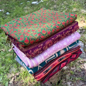 May include: A stack of colorful, folded textiles rests on a tree stump. The textiles display intricate patterns in shades of green, red, purple, pink, and teal. The stack is positioned outdoors, surrounded by grass and trees.