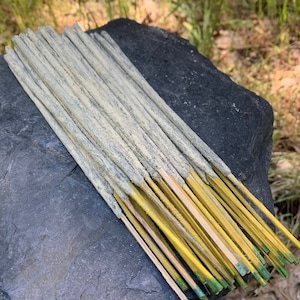 Puede incluir: Un conjunto de varillas de incienso, cada una con un cuerpo claro y texturizado y una base amarilla. Las puntas son de un verde apagado. El incienso descansa sobre una roca gris oscura y texturizada, con un fondo natural borroso.