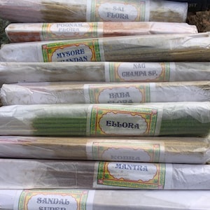 May include: A stack of incense sticks wrapped in white paper with colorful labels. The labels have names like "Saji Flora", "Poomam Flora", "Mysore Sandal", "Nag Champa SP", "Baba Flora", "Ellora", "Kobra Mantra", "Sandal Super", "Natural", and "Rudraksh".