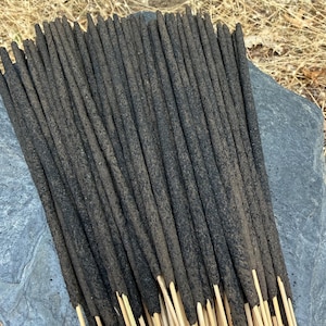 May include: A bundle of numerous incense sticks featuring dark grey, textured, cylindrical incense material coated onto thin, light-coloured wooden sticks. The incense sticks are arranged in a fan-like cluster, showing the uniform length and composition.