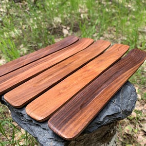 Könnte beinhalten: Fünf hölzerne Räucherstäbchenhalter in verschiedenen Brauntönen, von hell bis dunkel, sind auf einer grauen Steinoberfläche angeordnet. Die Halter sind rechteckig mit einer leichten Krümmung und einer Nut für Räucherstäbchen. Der Hintergrund zeigt grünes Gras und Blätter.