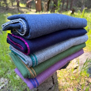 May include: A stack of folded, colorful wool scarves. The top scarf is a medium blue with a light gray border. Other scarves are navy, gray, green, and purple, each with a contrasting border in pink, teal, and gold.