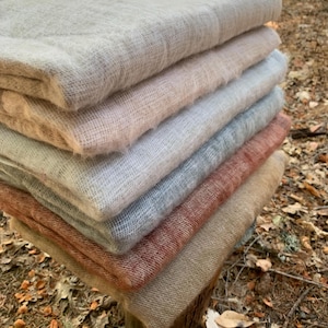May include: A stack of folded, soft blankets in various neutral colors, including beige, pink, gray, and rust. The blankets are made of a textured, woven material and are neatly arranged on a wooden surface, with a natural outdoor background.