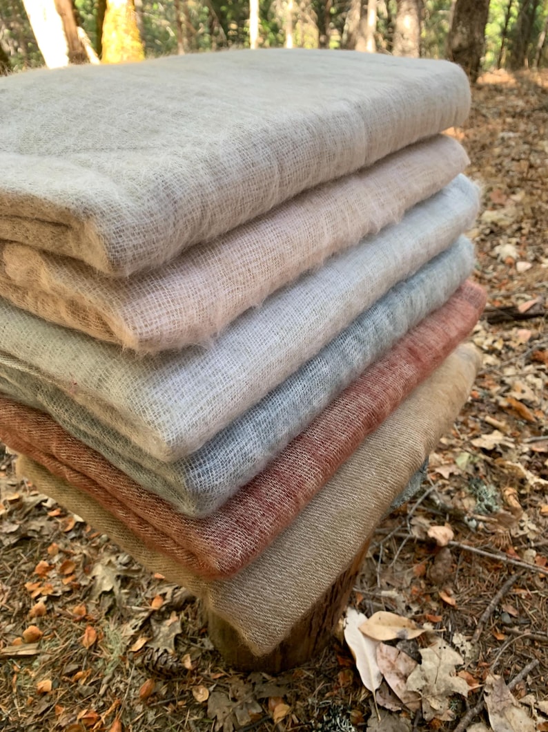 May include: A stack of folded, soft blankets in various neutral colors, including beige, pink, gray, and rust. The blankets are made of a textured, woven material and are neatly arranged on a wooden surface, with a natural outdoor background.