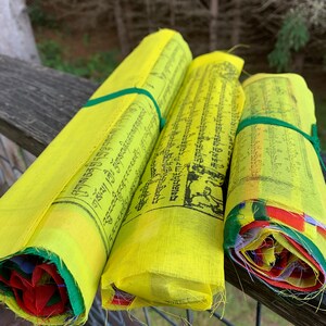 Peut inclure: Trois drapeaux de pri&egrave;re jaunes avec du texte noir et des liens verts sont enroul&eacute;s et empil&eacute;s sur une balustrade en bois. Les drapeaux sont li&eacute;s ensemble par un ruban vert. Les drapeaux sont devant un fond vert feuillu.