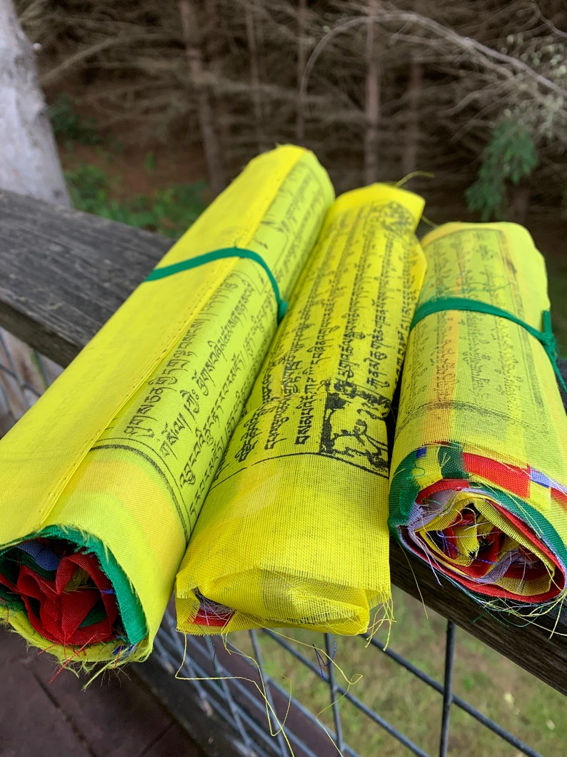 Peut inclure: Trois drapeaux de pri&egrave;re jaunes avec du texte noir et des liens verts sont enroul&eacute;s et empil&eacute;s sur une balustrade en bois. Les drapeaux sont li&eacute;s ensemble par un ruban vert. Les drapeaux sont devant un fond vert feuillu.