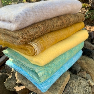 May include: A stack of five soft, woven scarves in various colors, including white, brown, yellow, and blue. The scarves are folded and stacked on top of each other, creating a colorful and textured display.