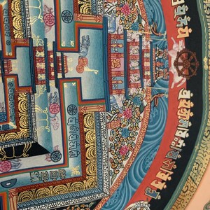 May include: A colorful mandala with intricate details, featuring a central square surrounded by a circular border. The mandala is painted in shades of blue, red, gold, and black, with floral and geometric patterns. The central square contains a smaller mandala with a wheel-like design.