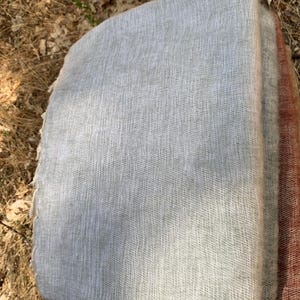 May include: A stack of folded, light gray woven fabric, possibly a blanket or throw. The fabric has a textured appearance and a soft, neutral color. A section of a reddish-brown fabric is visible on the right side of the stack.