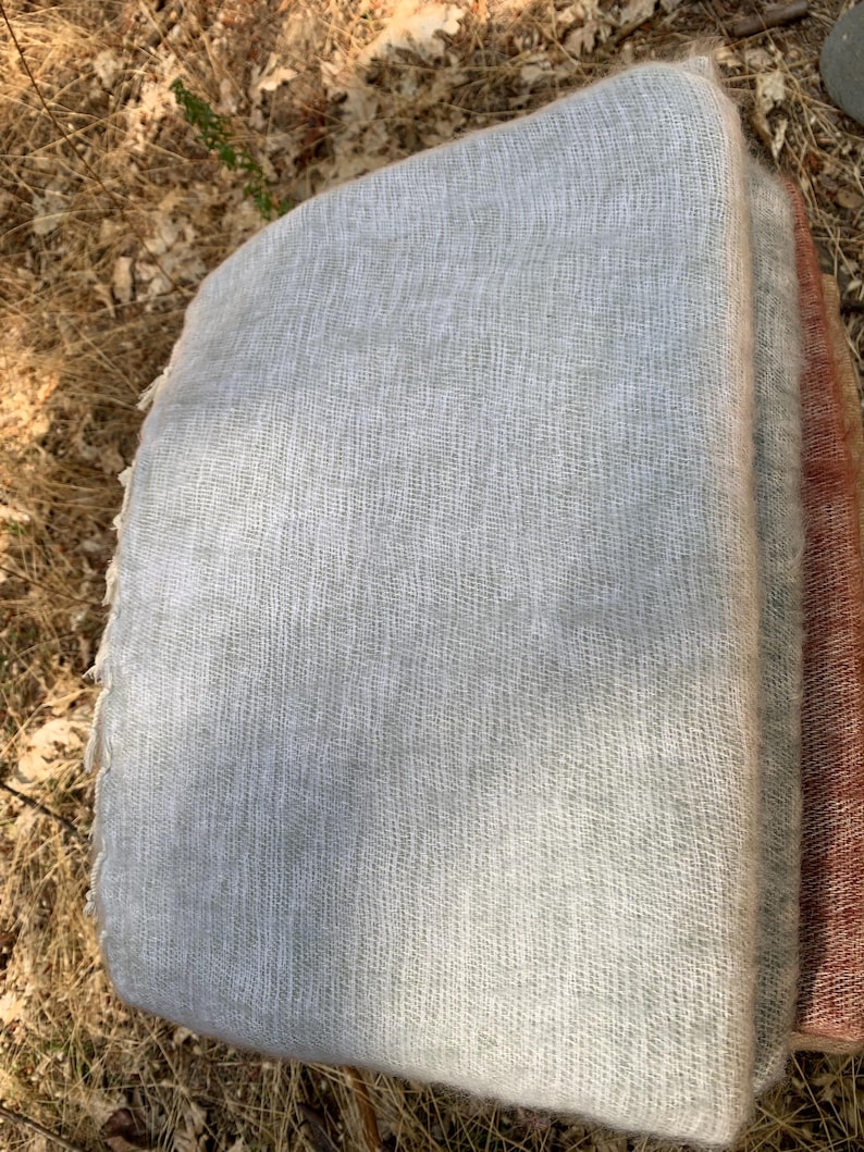 May include: A stack of folded, light gray woven fabric, possibly a blanket or throw. The fabric has a textured appearance and a soft, neutral color. A section of a reddish-brown fabric is visible on the right side of the stack.