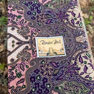 Può includere: Un pezzo di tessuto con un complesso motivo batik nelle tonalità del viola, rosa e crema con intricati motivi floreali e di foglie. Un'etichetta rettangolare è fissata al centro, riportante il testo Rimba Bali sopra un'illustrazione grafica.
