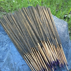 May include: A bundle of incense sticks with dark brown bodies and light brown tips. The sticks are arranged on a gray stone, with green grass in the background. The tips of the sticks are dyed purple.