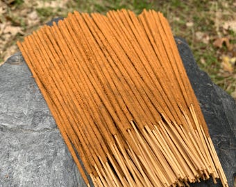 Puede incluir: Un conjunto de varillas de incienso de color marrón cálido y terroso. El incienso está dispuesto sobre una superficie de piedra gris oscuro, con un fondo borroso de hojas verdes y marrones. Las varillas tienen un aspecto texturizado y están agrupadas.