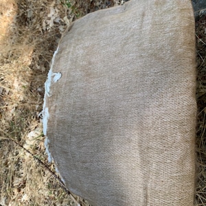 May include: A rolled-up, tan-colored fabric with a textured weave. The edge of the fabric is frayed, revealing a white interior. The fabric is lying on a bed of dry grass and leaves, suggesting an outdoor setting.