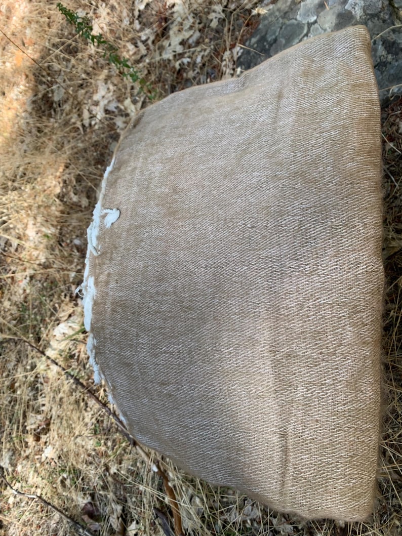 May include: A rolled-up, tan-colored fabric with a textured weave. The edge of the fabric is frayed, revealing a white interior. The fabric is lying on a bed of dry grass and leaves, suggesting an outdoor setting.
