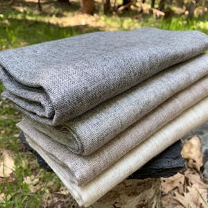 May include: A pile of folded fabric in shades of grey and beige. The fabrics are neatly arranged, displaying a variety of textures and tones. The image is taken outdoors, with a blurred backdrop of trees and greenery.