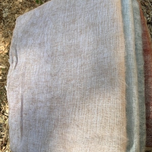 May include: A stack of woven fabric in various neutral colors. The top layer is a light beige, followed by a pale blue-gray, and a muted reddish-brown. The fabric has a loose, open weave, suggesting a lightweight texture.
