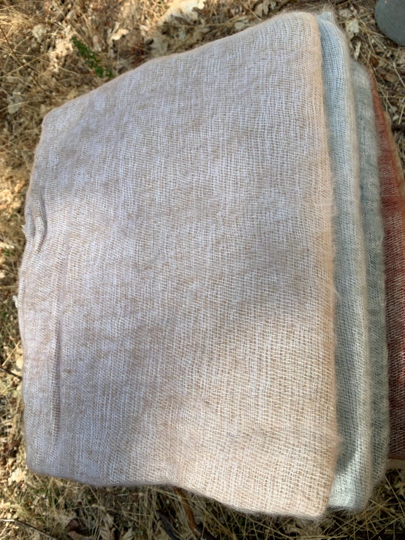 May include: A stack of woven fabric in various neutral colors. The top layer is a light beige, followed by a pale blue-gray, and a muted reddish-brown. The fabric has a loose, open weave, suggesting a lightweight texture.