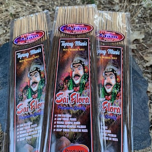 May include: Three packages of incense sticks. Each package features a label with a cartoon illustration of a smiling figure in traditional Balinese attire. The labels also display the text "Topeng Manis" and "Sai Flora Premium Quality."