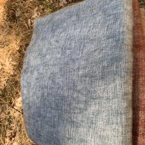 May include: A folded, light blue scarf with a soft, open weave texture. The scarf has a gradient effect, transitioning from a darker blue to a lighter, almost white color. A section of a rust-colored scarf is visible on the right.