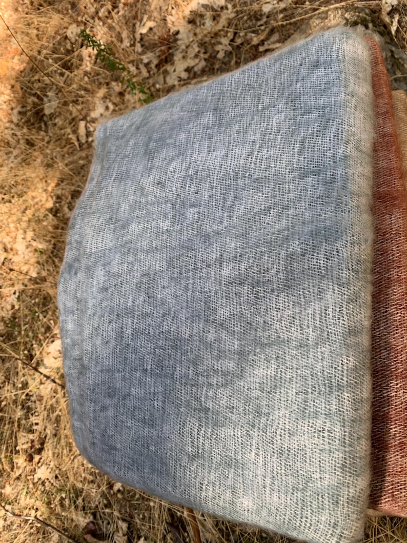 May include: A folded, light blue scarf with a soft, open weave texture. The scarf has a gradient effect, transitioning from a darker blue to a lighter, almost white color. A section of a rust-colored scarf is visible on the right.