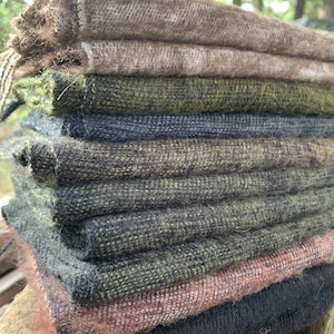May include: A stack of woven fabric in various earthy tones. The colors range from light brown and olive green to darker shades of gray and black. The fabric has a textured appearance, suggesting a natural fiber like wool or linen.