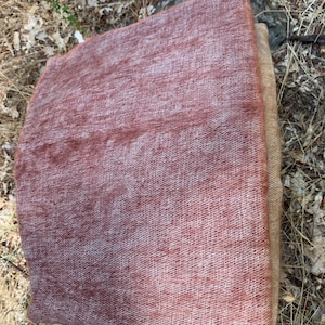 May include: A rectangular, woven textile in shades of dusty rose and tan. The fabric has a textured appearance, with visible threads creating a slightly uneven surface. The item is folded, revealing a layered construction.