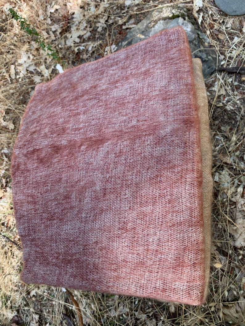 May include: A rectangular, woven textile in shades of dusty rose and tan. The fabric has a textured appearance, with visible threads creating a slightly uneven surface. The item is folded, revealing a layered construction.