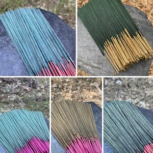 May include: Bundles of incense sticks in various colors, including teal with red tips, green with gold tips, and green with pink tips. The incense is arranged on a dark gray stone surface, with a blurred natural background.