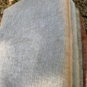 May include: A stack of soft, woven scarves in shades of gray, beige, and muted red. The fabric has a delicate, open weave, suggesting a lightweight texture. The scarves are neatly folded, showcasing their layered colors.