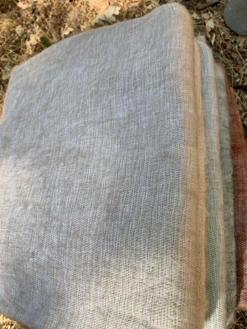 May include: A stack of soft, woven scarves in shades of gray, beige, and muted red. The fabric has a delicate, open weave, suggesting a lightweight texture. The scarves are neatly folded, showcasing their layered colors.