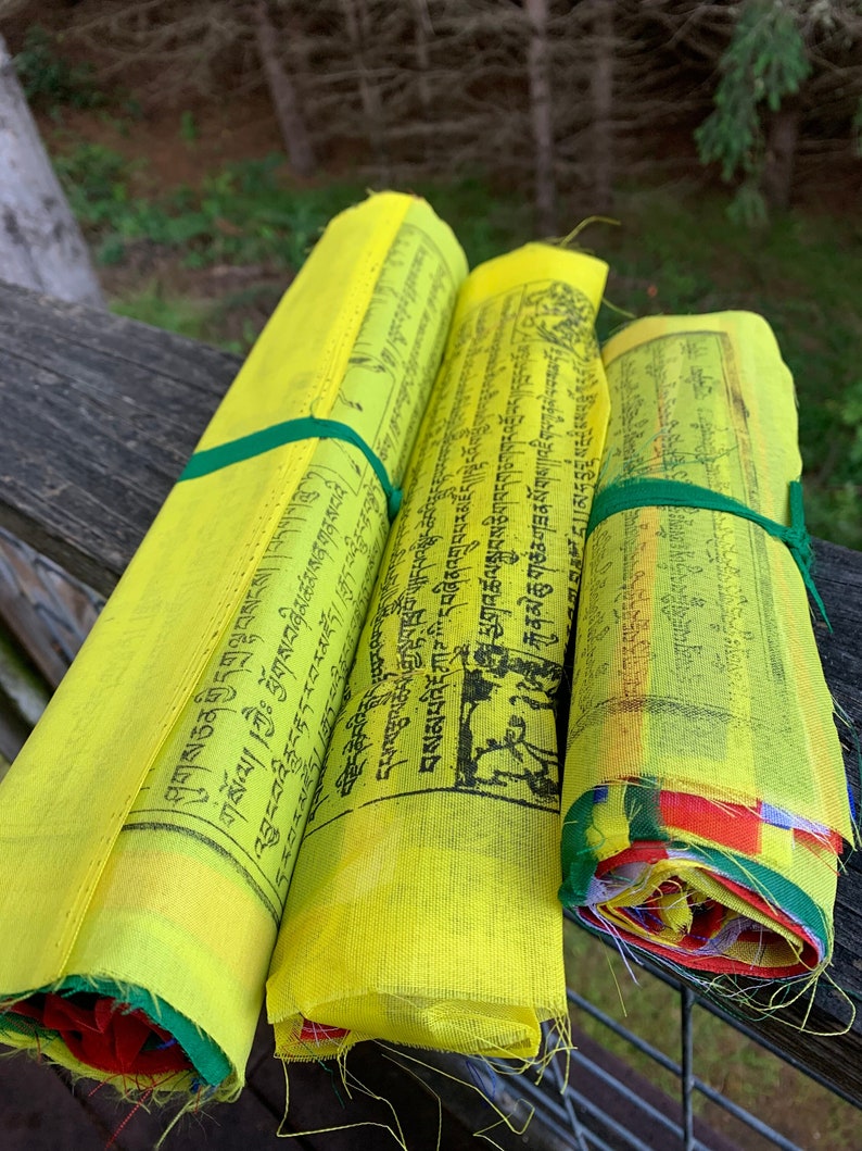 Peut inclure: Trois drapeaux de pri&egrave;re jaunes enroul&eacute;s avec du texte noir et des bordures color&eacute;es. Les drapeaux sont li&eacute;s ensemble avec une ficelle verte.