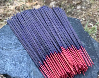 May include: A bundle of incense sticks with purple shafts and red tips rests on a gray stone. The incense is arranged in a fan shape, with the red tips creating a vibrant contrast against the purple.