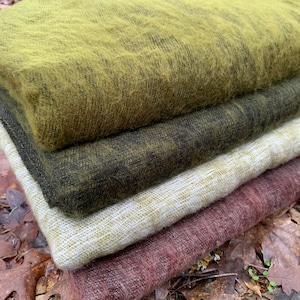 May include: A stack of four woven blankets in different colors and textures. The top blanket is a bright green, the second is a dark green, the third is a light beige, and the bottom is a deep burgundy.