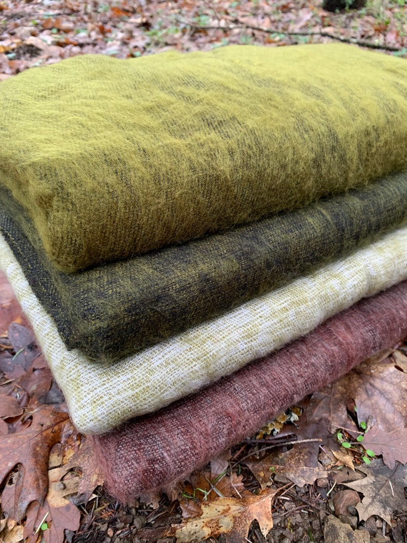May include: A stack of four woven blankets in different colors and textures. The top blanket is a bright green, the second is a dark green, the third is a light beige, and the bottom is a deep burgundy.