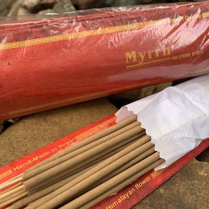 Puede incluir: Un paquete de inciensos de mirra, hechos a mano en Nepal y decorados con flores del Himalaya. El embalaje es rojo con letras doradas y un borde dorado. Los inciensos son marrones y están agrupados.
