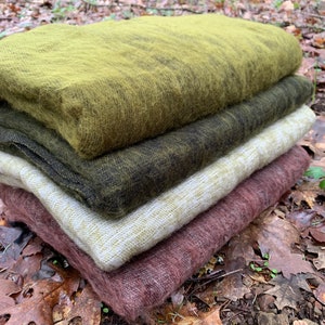 May include: A stack of four soft, fuzzy blankets in various shades of green, brown, and pink. The blankets are folded and arranged on a bed of fallen leaves.