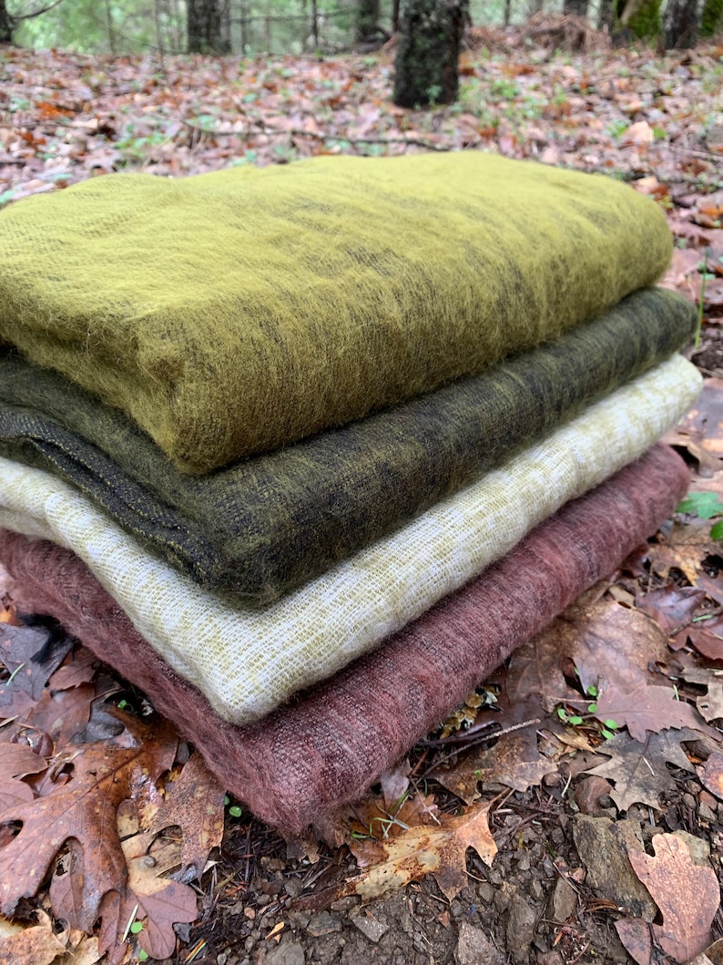 May include: A stack of four soft, fuzzy blankets in various shades of green, brown, and pink. The blankets are folded and arranged on a bed of fallen leaves.