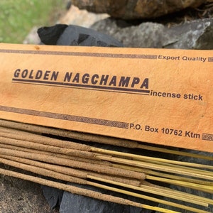 Puede incluir: Una caja de inciensos Golden Nag Champa. La caja es marrón con letras doradas y un borde. El texto en la caja dice "Golden Nag Champa incense stick" y "Export Quality". La caja también tiene un número de apartado de correos y una ubicación.
