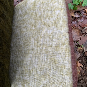 May include: A green and white woven throw blanket with a soft, fuzzy texture. The blanket is folded and lying on a bed of brown leaves.
