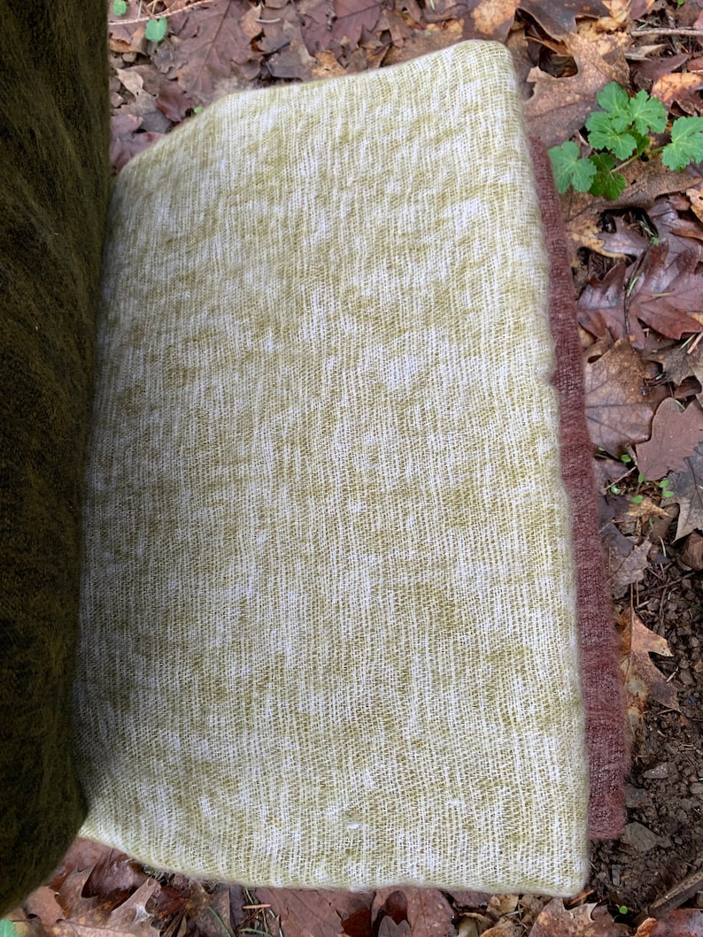 May include: A green and white woven throw blanket with a soft, fuzzy texture. The blanket is folded and lying on a bed of brown leaves.