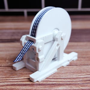 May include: A white ribbon dispenser with a spool of blue and white striped ribbon. The dispenser is made of white plastic and sits on a wooden surface. The ribbon is partially unwound.