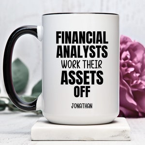 Może przedstawiać: Biały kubek ceramiczny z czarnym uchwytem i czarnym rantem. Kubek ma czarny napis "Financial Analysts Work Their Assets Off" i imię "Jonathan" pod napisem.