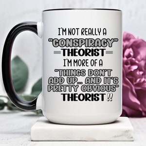 Puede incluir: Taza de café de cerámica blanca con asa negra. La taza tiene un borde negro y presenta el texto "I'm not really a "Conspiracy" Theorist= I'm more of a "Things don't add up... and it's pretty obvious" Theorist!!"