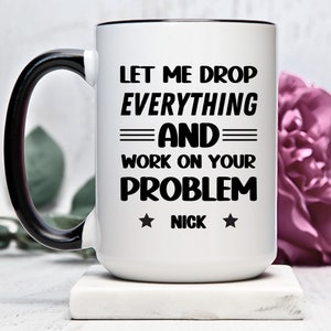 Puede incluir: Taza de cerámica blanca con asa y borde negros. La taza tiene texto negro que dice "Let me drop everything and work on your problem" con una estrella a cada lado del nombre "Nick".
