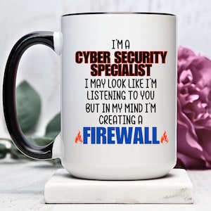 May include: White ceramic coffee mug with a black handle and black rim. The mug has a humorous saying about cyber security specialists: "I'm a Cyber Security Specialist. I may look like I'm listening to you, but in my mind I'm creating a firewall."